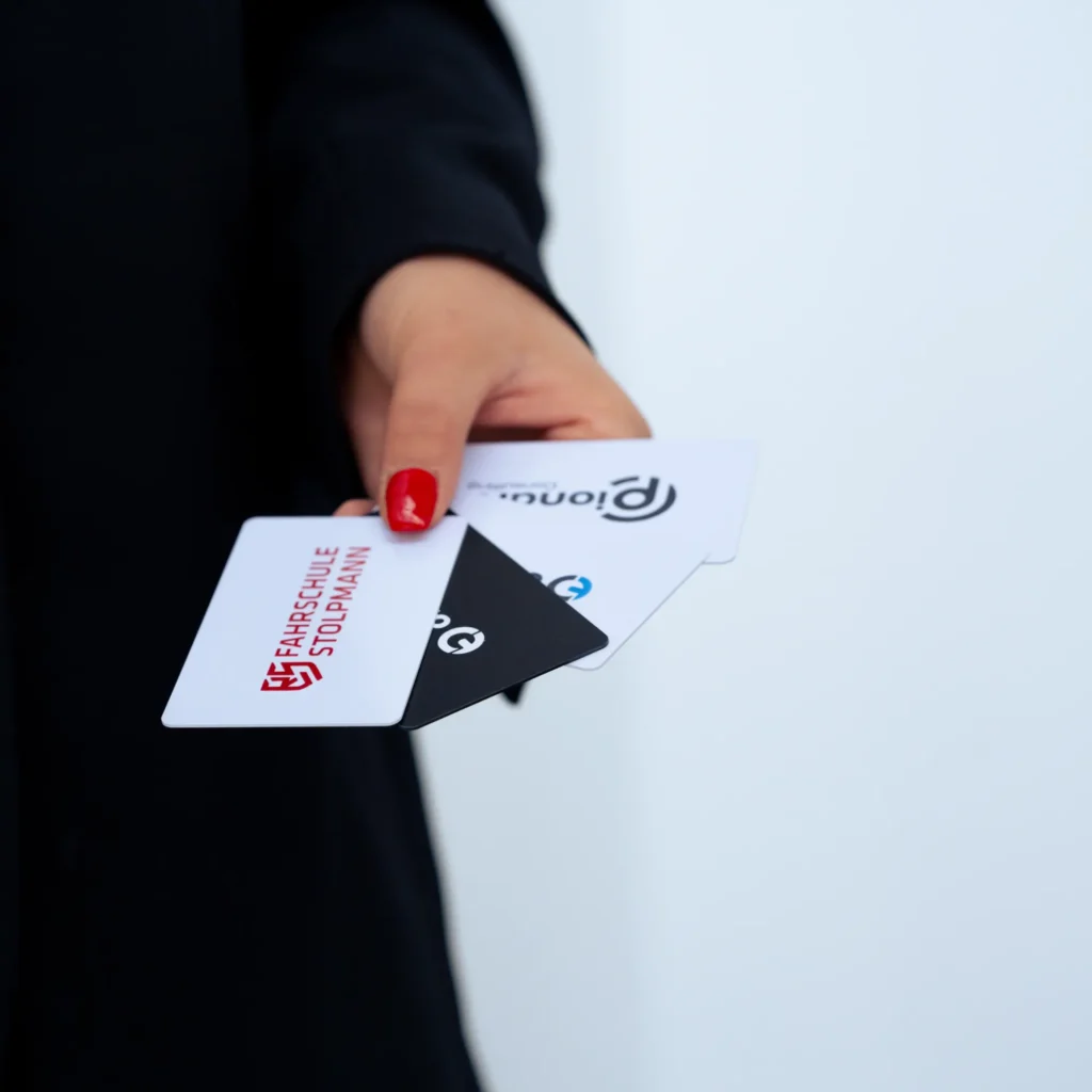 Eine junge Frau mit rot lackierten Fingernägeln hält vier NFC Visitenkarten aufgefächert in der Hand. Man sieht die Logos von onlytap, der Fahrschule Stolpmann und der PIONARA Consulting.